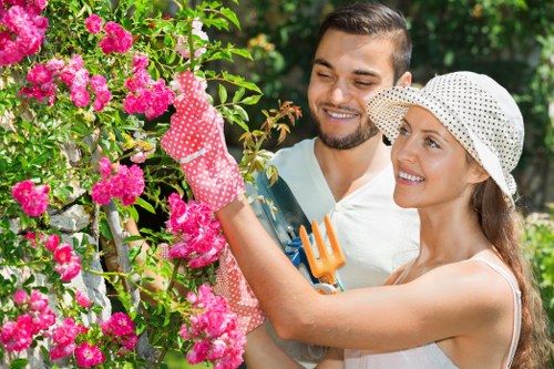 A woman and a man are working together in a lush garden, tending to a vibrant flowering shrub with bright pink blooms. The woman, wearing a wide-brimmed, white polka-dot sunhat and gardening gloves, is pruning the plant with a small pair of garden scissors. The man, with short dark hair and a trimmed beard, stands closely beside her, smiling and holding a small orange gardening fork. The garden features a well-maintained lawn in the foreground, with a backdrop of green trees and shrubs, suggesting a landscaped outdoor space typical of homes in Harrow. The scene is illuminated by natural sunlight, indicating fair weather, and the garden is surrounded by a mix of flowering plants, hedges, and a paved pathway. This image reflects professional gardening activities that could be offered by Gardening Services Harrow, showcasing meticulous plant care within a tidy, inviting garden environment near postcode HA postcode area.